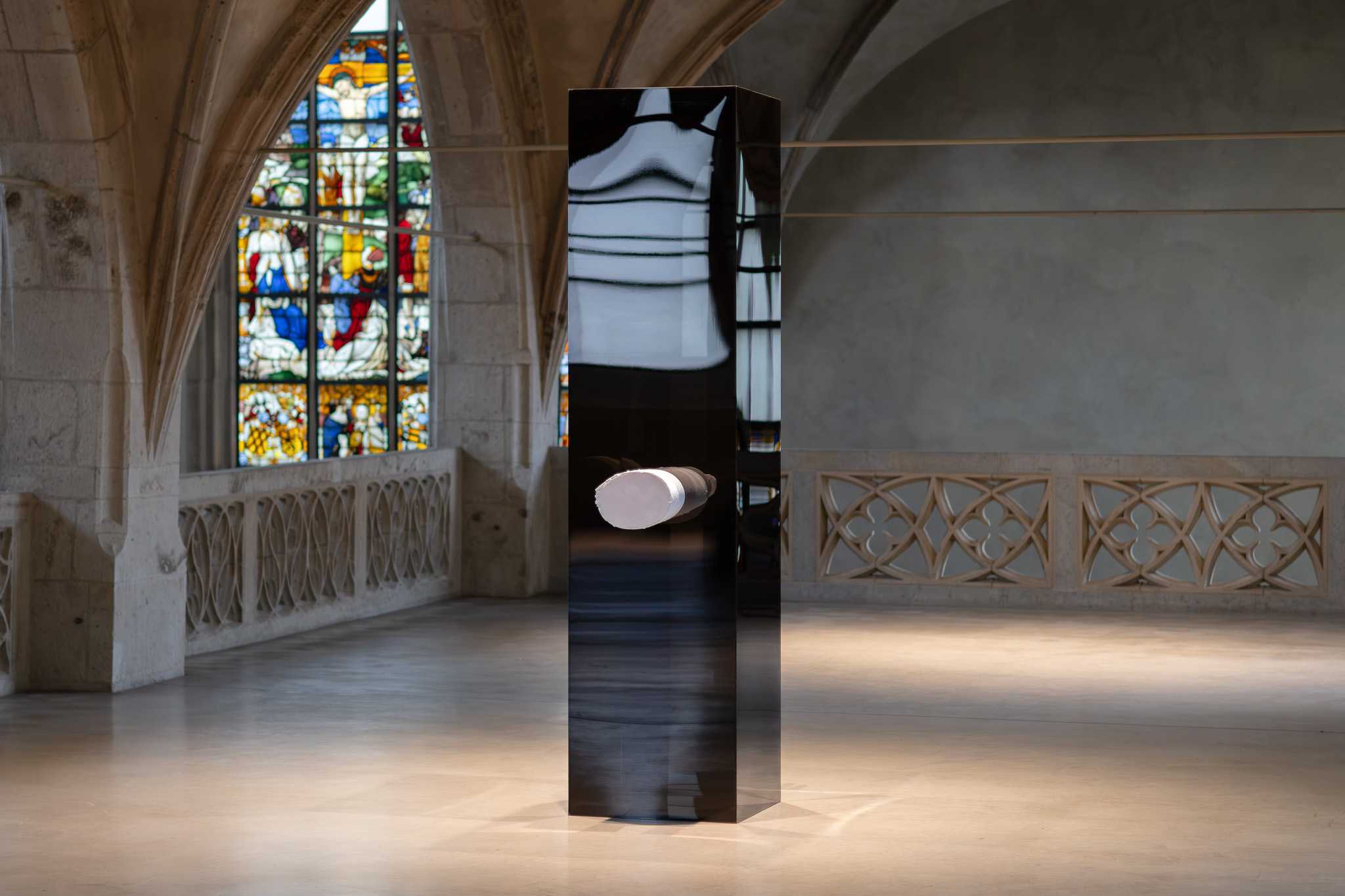 A tall black plinth in a vaulted church interior, a stained-glass window behind it; a white 3D-printed arm reliquary projects from its front face.