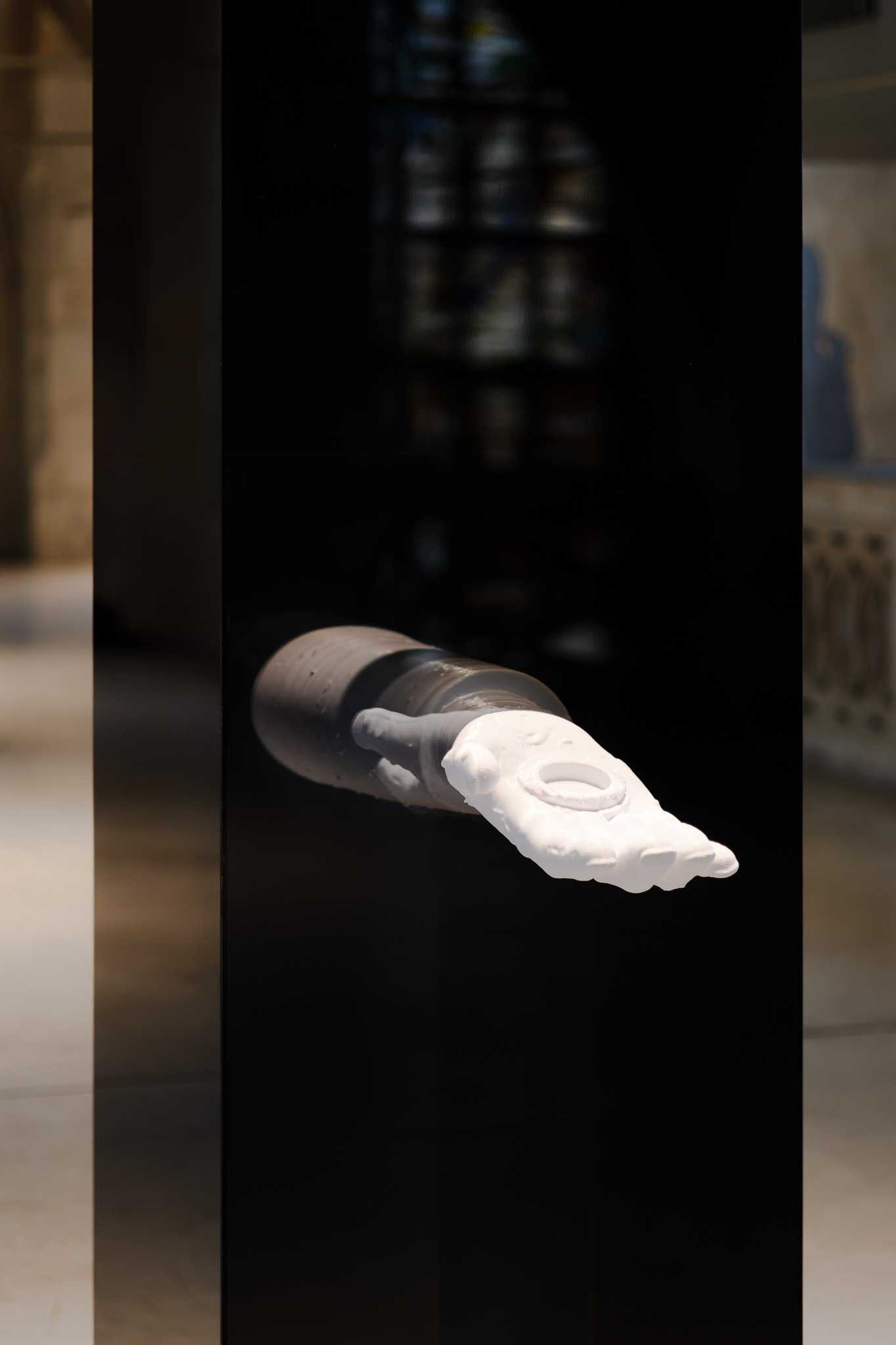 Close view of the white 3D-printed arm reliquary emerging from the black reflective plinth.