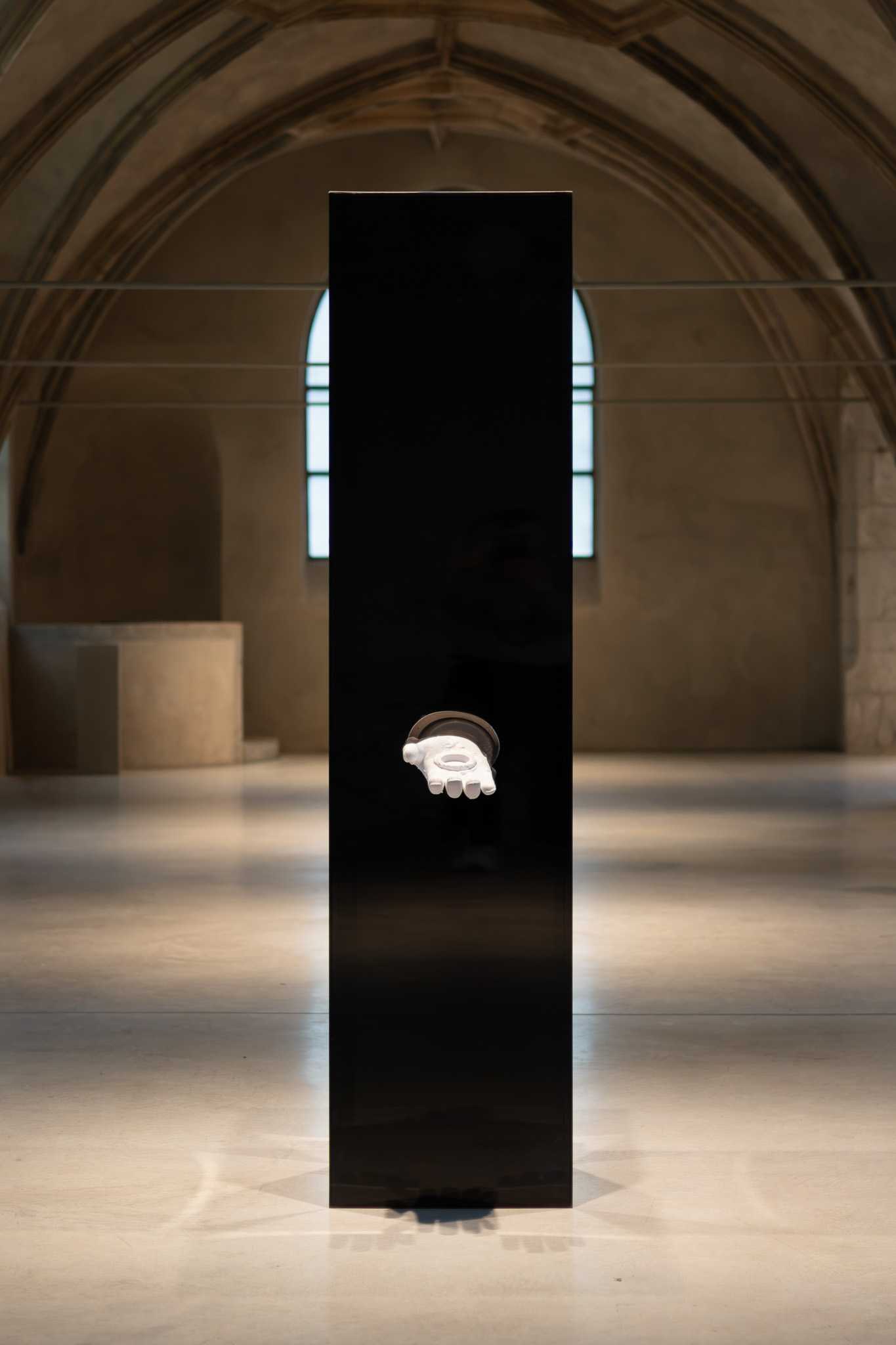 The black plinth seen head-on beneath the vaulting, an arched window behind it, the 3D-printed arm reliquary held out from its face.