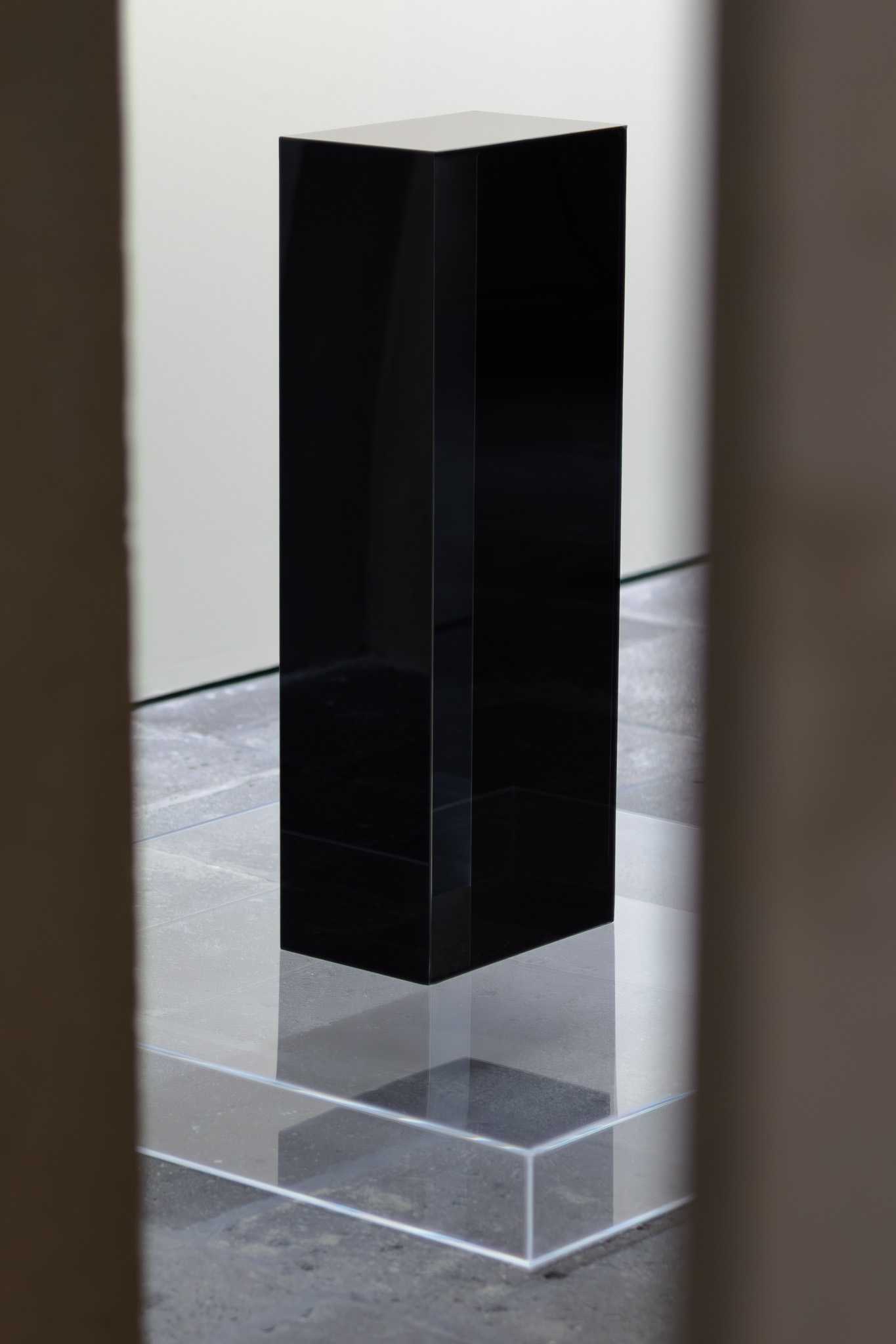 A black glossy column standing on a clear acrylic base plate, seen through a narrow opening.