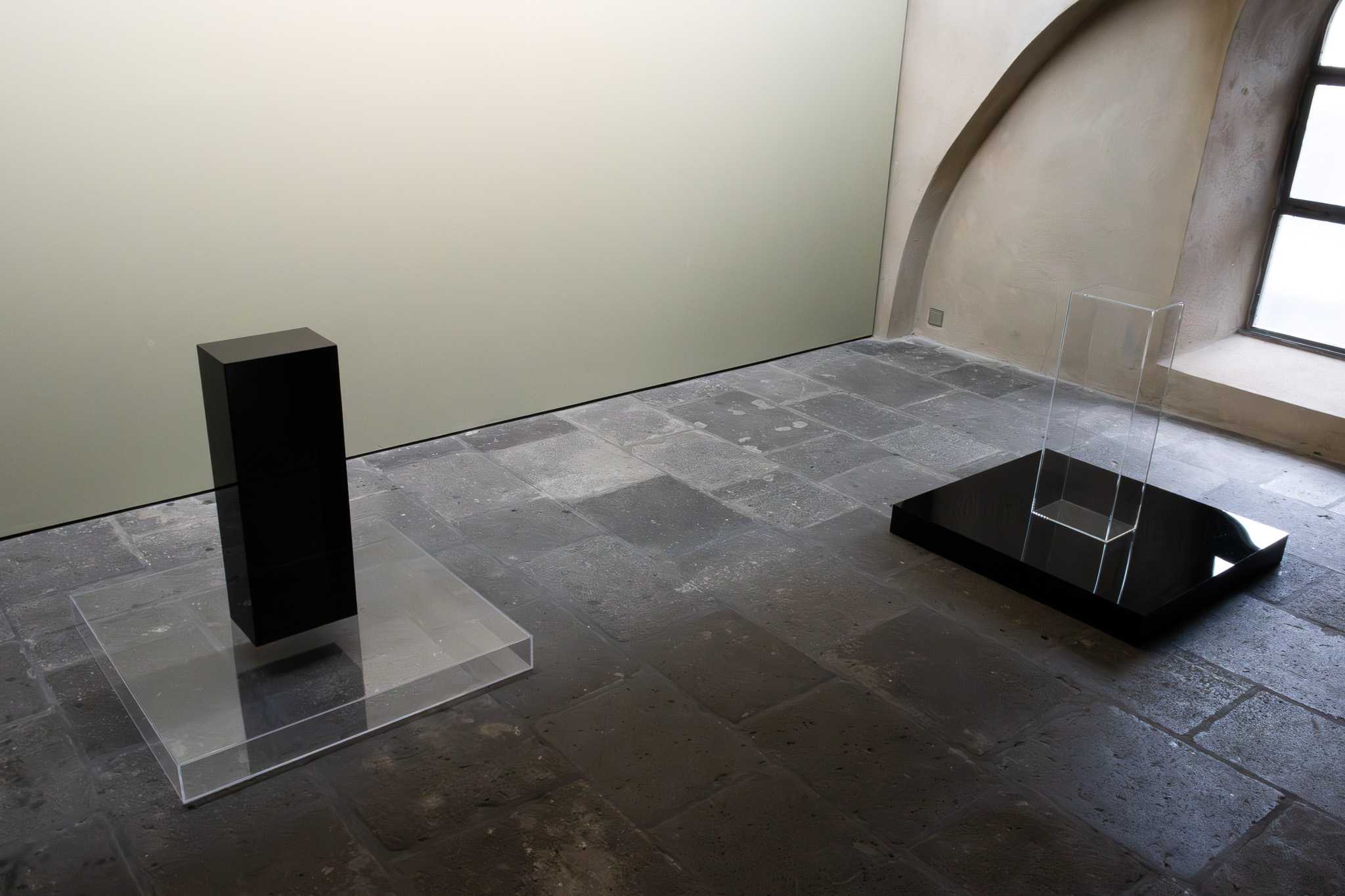 Room view with both works: at left a black column on a clear acrylic plate, at right a clear acrylic case on a black plate.