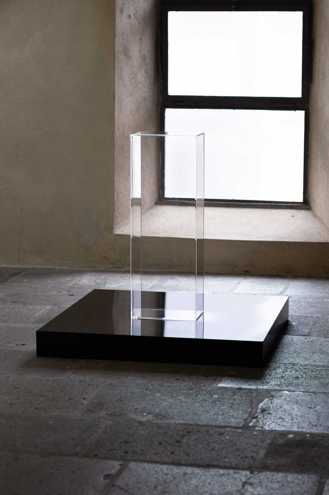 A clear acrylic case standing on a low black plinth, backlit by a window.