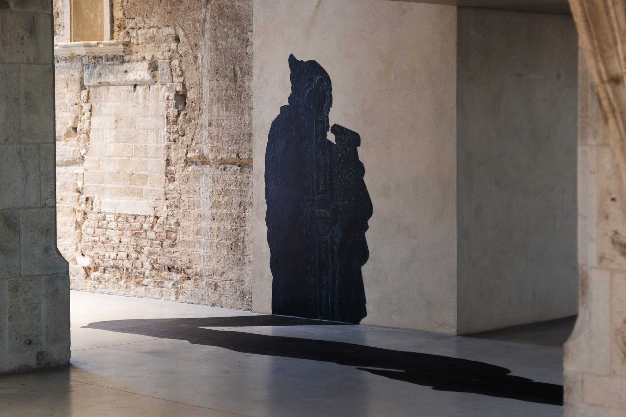 A black silhouette of one of the Cologne city hall prophets, holding a scroll, applied to a plaster pier; its flat shadow extends across the polished floor.