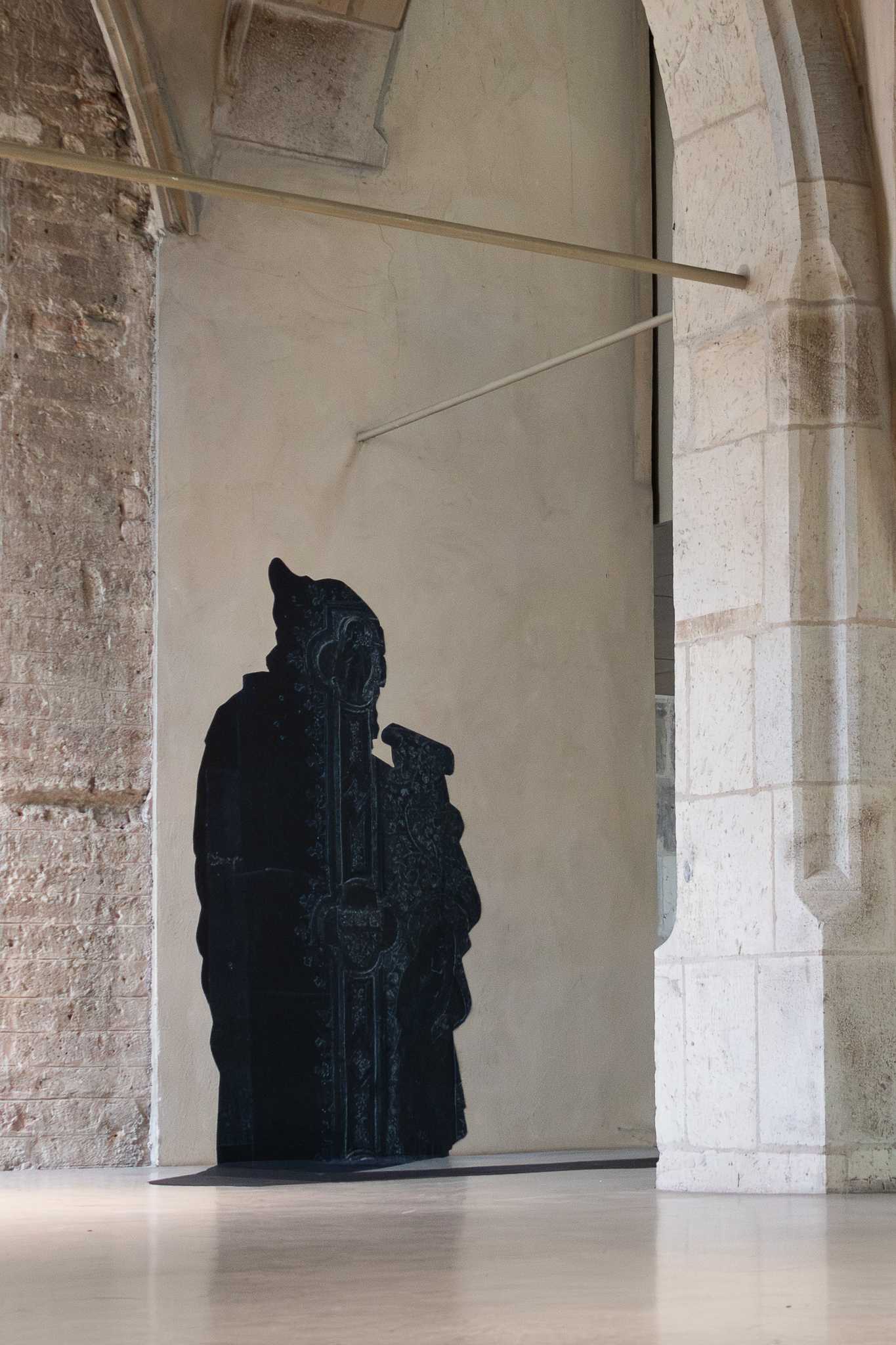 The prophet at full height against a plastered bay of wall, rough brick to the left and a pale stone pier to the right, a slender hanging rail crossing above and a low shadow pooling at its foot.